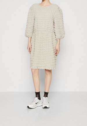 Struktureret beige kjole med trekvart pufærmer, elastisk talje og løs pasform, parret med hvide sneakers og sorte gennemsigtige strømper.