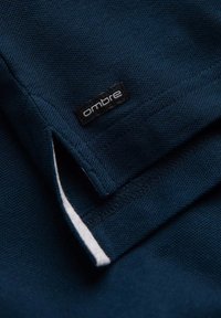 Tessuto blu navy con texture e un'etichetta "ombre" ben visibile. Presenta un bordo pulito e rifinito con cura e un dettaglio a patta.