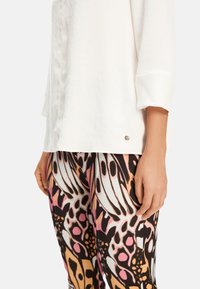 Weiße Bluse mit strukturierter Oberfläche und umgeschlagenen Ärmeln, kombiniert mit engen Leggings, die mit einem bunten Schmetterlingsflügel-Print in Pink, Schwarz und Orange versehen sind.