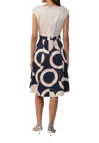Marineblauwe en roomwitte jurk met een cirkelvormig patroon, korte mouwen, getailleerde taille en een rok tot op de knie. Achterste ritsluiting. Zilveren schoeisel zichtbaar.