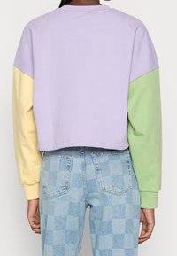 Persona vistiendo una sudadera recortada con cuerpo lavanda, manga izquierda amarilla y manga derecha verde, combinada con jeans a cuadros azul claro.