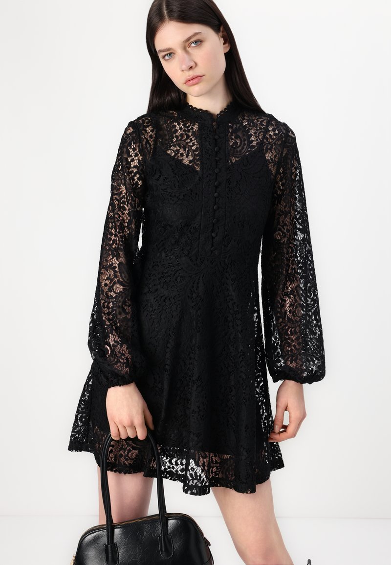 Schwarzes Spitzenkleid mit langen, gepufften Ärmeln; mit hohem Ausschnitt und Knopfdetails. Getragen mit einer schwarzen Handtasche, die komplizierte Muster zeigt.