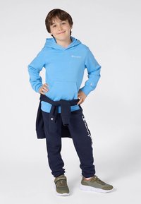 Camisola com capuz azul-claro com bolso frontal e logótipo, combinada com calças de fato de treino azul-marinho com marca branca, e ténis verdes com solas brancas.