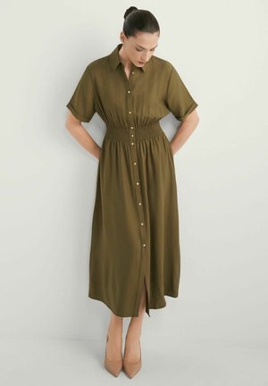 Kobieta z ciemnymi włosami upiętymi w kok, ubrana w oliwkowozieloną sukienkę midi na guziki z krótkimi rękawami i beżowymi, spiczastymi szpilkami, stojąca na tle białej ściany.