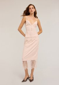 Vestido de encaje rosa pálido con un corsé ajustado, escote profundo y un ribete de encaje en el dobladillo. Las tirantes delgados y un sutil detalle de botones añaden elegancia.