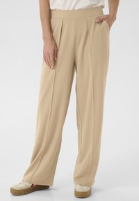 Beige wide-leg trousers with pleats, made of a smooth fabric. Features front pockets and paired with light-toned sneakers.