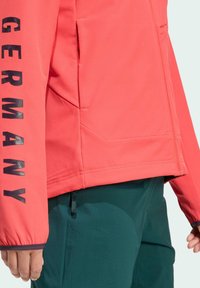 Koraalrode jas met zwarte tekst "GERMANY" op de mouw, met zacht materiaal, zakken met rits en een aangesloten ontwerp met elastische manchetten.