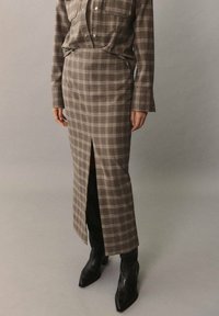 Longue jupe à carreaux en tissu marron et gris, avec une fente avant ; associée à des bottines noires ajustées et une chemise à carreaux assortie.