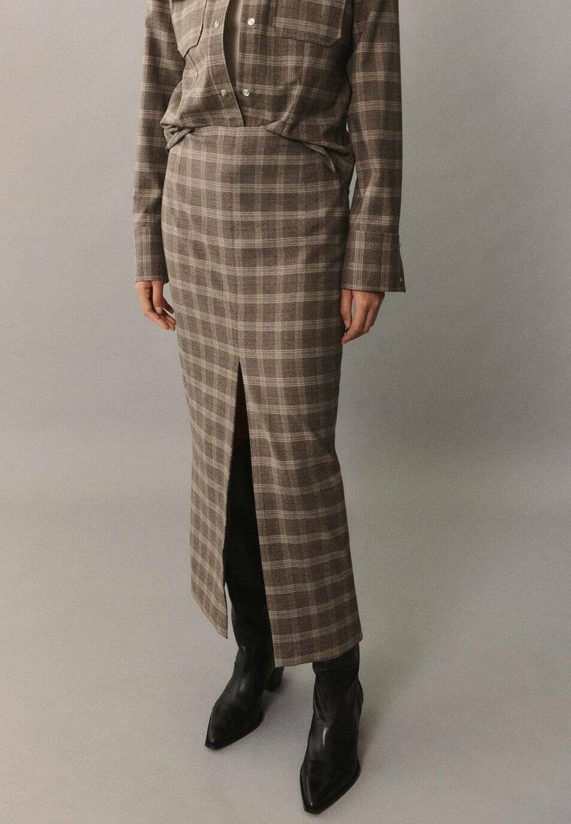 Longue jupe à carreaux en tissu marron et gris, avec une fente avant ; associée à des bottines noires ajustées et une chemise à carreaux assortie.