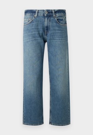 Vrouw staand met handen in de zakken, draagt een wit topje met korte mouwen, blauwe cropped jeans, witte sokken en zwarte loafers op een witte achtergrond.