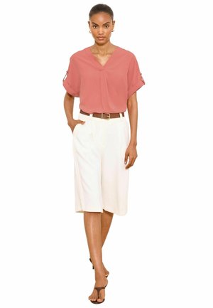 Kobieta ubrana w łososiową bluzkę z dekoltem w kształcie litery V z podwiniętymi krótkimi rękawami, białe szorty do kolan z paskiem i czarne sandały, stojąca z jedną ręką w kieszeni.