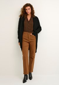 Schwarze Strickjacke, gemusterte braune Bluse mit kleinen roten Punkten, braune gerade Hosen und schwarze Stiefeletten.