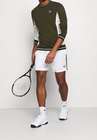 Camisa de manga larga verde oliva con paneles laterales blancos, pantalones cortos blancos y zapatillas blancas. Sosteniendo una raqueta de tenis negra.