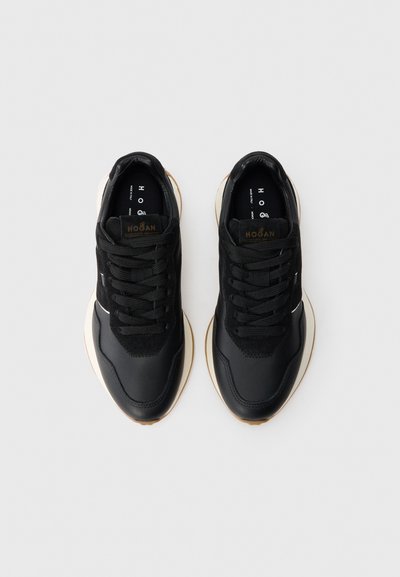 Adidași sport negri cu un exterior din piele netedă și sueded, având șireturi negre, o talpă intermediară albă și o talpă din cauciuc de tip gumă.