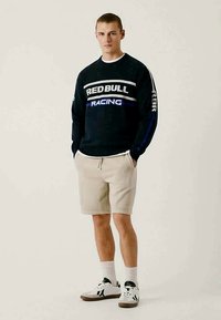 Un giovane uomo è in piedi indossando una felpa nera della Red Bull Racing, pantaloni beige, calze bianche e sneakers bianche con dettagli neri.