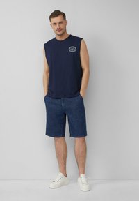 Navy ärmelloses Shirt mit runder Logo, kombiniert mit knielangen dunklen Jeansshorts und weißen Sneakers. Glatter Stoff, lässiger Schnitt.