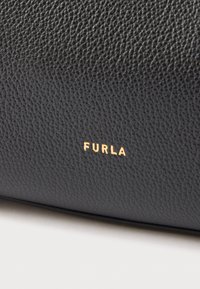 Melnas ādas soma ar strukturētu virsmu. Uz tās apakšējā labajā stūrī atrodas zelta uzspiests "FURLA" logotips. Kompakta taisnstūrveida forma.