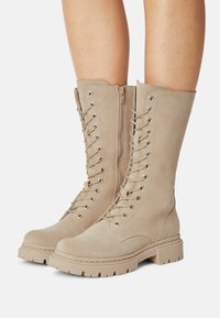 Beige suede knee-high boots with lace-up front, metal eyelets, and a side zipper; chunky rubber sole with traction grip pattern.