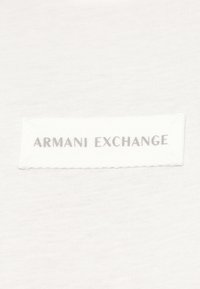 White fabric with a stitched rectangular patch displaying the text "ARMANI EXCHANGE" in silver letters.