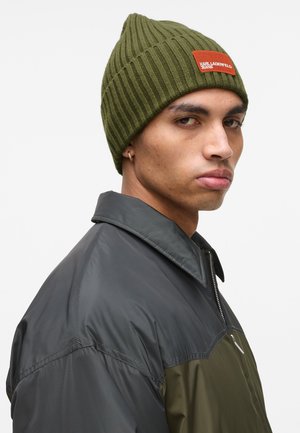 Olive green ribbed knit beanie with a pointed top and an orange rectangular logo patch. Worn by a person in a dark, zippered jacket.