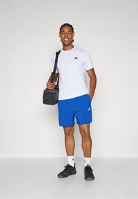 Tricou alb cu maneci scurte, cu logo negru, pantaloni scurți albaștri, adidași negri și o geantă gri. Material neted cu o croială lejeră și accente.