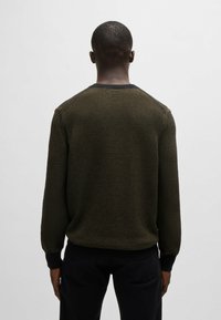 Maglione lavorato a maglia in oliva scuro con collo rotondo a coste e motivo testurizzato. Maniche e orlo aderenti, indossato sopra pantaloni neri.