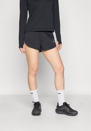 Personne portant un haut à manches longues noir, un short de sport noir, des chaussettes blanches Nike crew et des chaussures de running Nike noires, debout sur un sol gris.