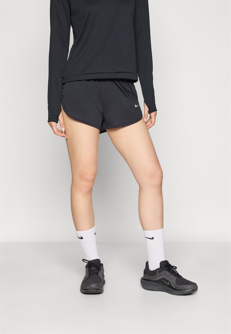 Personne portant un haut à manches longues noir, un short de sport noir, des chaussettes blanches Nike crew et des chaussures de running Nike noires, debout sur un sol gris.