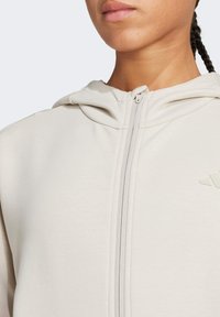 Light grey zip-up hoodie with a textured finish, featuring a hood and minimalistic branding on the chest. Zipper and seams are visible.