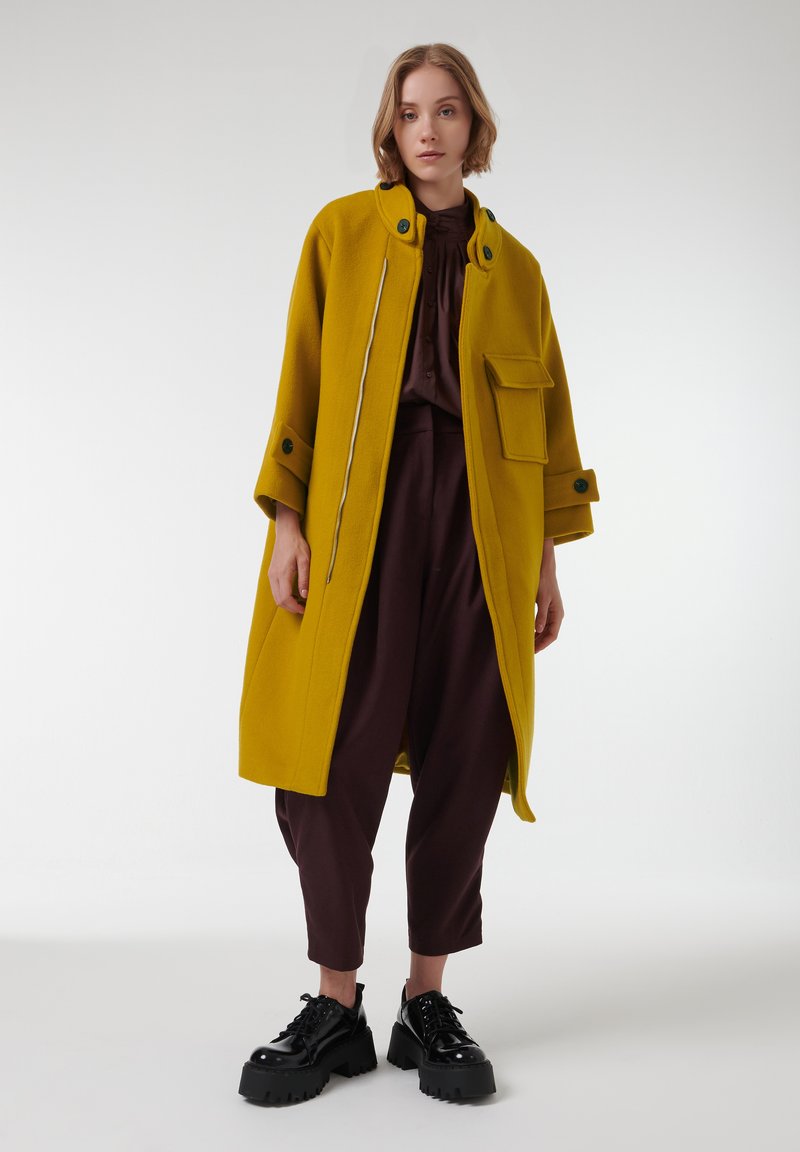 Abrigo de lana amarillo con un cuello alto, cremallera en la parte delantera y un gran bolsillo. Combinado con una blusa y pantalones marrón oscuro, y zapatos negros robustos.