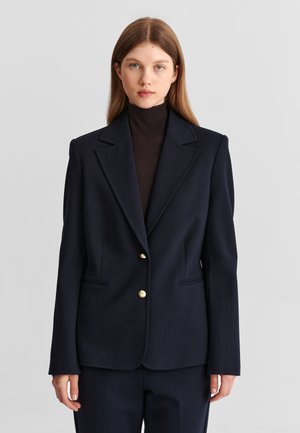 Jeune femme aux cheveux lisses châtain clair porte un blazer bleu marine foncé sur un col roulé noir devant un fond gris clair uni.