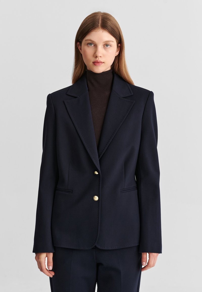 Jeune femme aux cheveux lisses châtain clair porte un blazer bleu marine foncé sur un col roulé noir devant un fond gris clair uni.