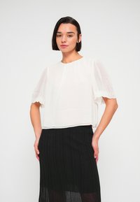 Femme aux cheveux courts et foncés portant un chemisier blanc à manches en dentelle et une jupe plissée noire, debout les mains sur les hanches.