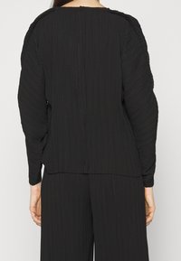 Svart plisserad topp med rund halsringning, långa ärmar och strukturerad tyg. Bakdelen har en rak söm och sitter något löst.