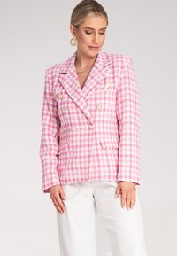 Dwurzędowa różowo-biała marynarka w kratę houndstooth z złotymi guzikami, z klapami w kształcie notcha i bocznymi kieszeniami, noszona na białych spodniach.