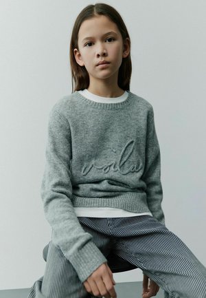 Grauer Strickpullover mit gerippten Bündchen, versehen mit aufgesticktem "voilà"-Text, getragen über einem weißen Hemd und kombiniert mit gestreiften Hosen.