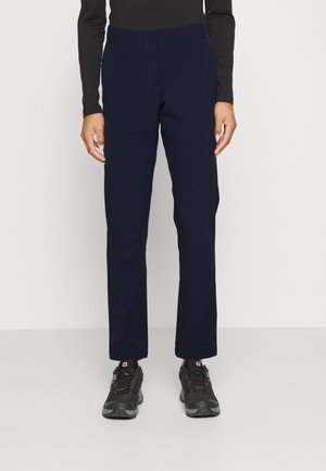 Person iført navy straight-leg bukser, sort langærmet top og sorte snøresports-sko, der står mod en enkel baggrund.