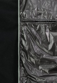 Schwarzer Fleece-Stoff auf der linken Seite kontrastiert mit glänzendem, zerknittertem, dunkelgrauem wasserdichtem Material auf der rechten Seite, getrennt durch eine schmale graue Naht.