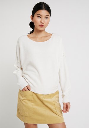 Maglione bianco oversize con un ampio scollo a barca, trama morbida, abbinato a una mini gonna in velluto a coste color senape con tasche frontali.