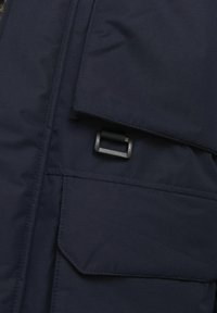 Marineblauwe geïsoleerde jas van stof met een grote klepzak en verstelbare riemhardware. Glad van structuur met stiksels.