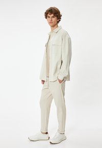 Camisa de corte relajado color beige claro con dos bolsillos en el pecho, llevada sobre una blusa beige y pantalones tobilleros a juego. Zapatillas deportivas blancas.