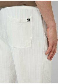 Weiße gestreifte Hose aus einem weichen Stoff, mit einer Gesäßtasche und einem Markenschild. Die Textur hat ein Fischgrat-Muster.