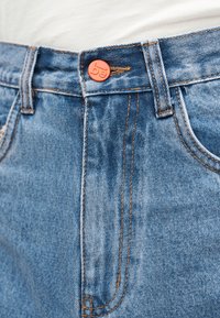 Ζώνη από μπλε τζιν με πορτοκαλί κουμπί και ορατή ραφή, φορεμένη με λευκό πουκάμισο στριμωγμένο μέσα.