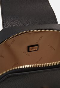Innenraum einer schwarzen Lederhandtasche mit strukturierter Oberfläche, einem goldfarbenen Reißverschluss, beige gefüttert mit wiederholtem "GUESS"-Logo und einem schwarzen Etikett mit goldenen Akzenten.
