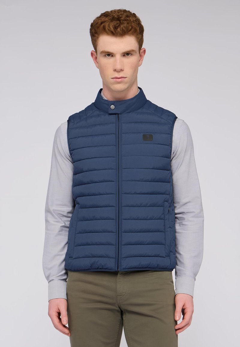 Námořnická puffer vesta s stojáčkem, vodorovným prošíváním, předním zipem a černou logovou nášivkou. Nositá přes světle šedou košili s dlouhým rukávem.