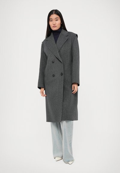 Cappotto grigio doppiopetto in misto lana testurizzata, con grandi rever a punta e due bottoni neri, indossato su un dolcevita blu navy e jeans blu.