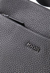 Bolso de cuero texturizado negro con un bolsillo con cremallera y un logo en relieve de "JOOP!" en la superficie.