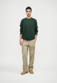 Mørkegrøn tekstureret sweater, afslappet pasform, ribbet krave. Lys tan cargo bukser med sidelommer. Brune sko med lav profil.