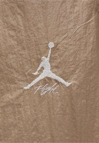 Tan tyg med en rynkad struktur har en vit broderad Jumpman-logga och ordet "Flight" under i kursiv stil.