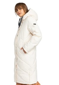 Manteau doudoune blanc avec capuche, présentant un design matelassé diagonal, des poches latérales et un patch logo noir sur la manche. Tissu doux et texturé.